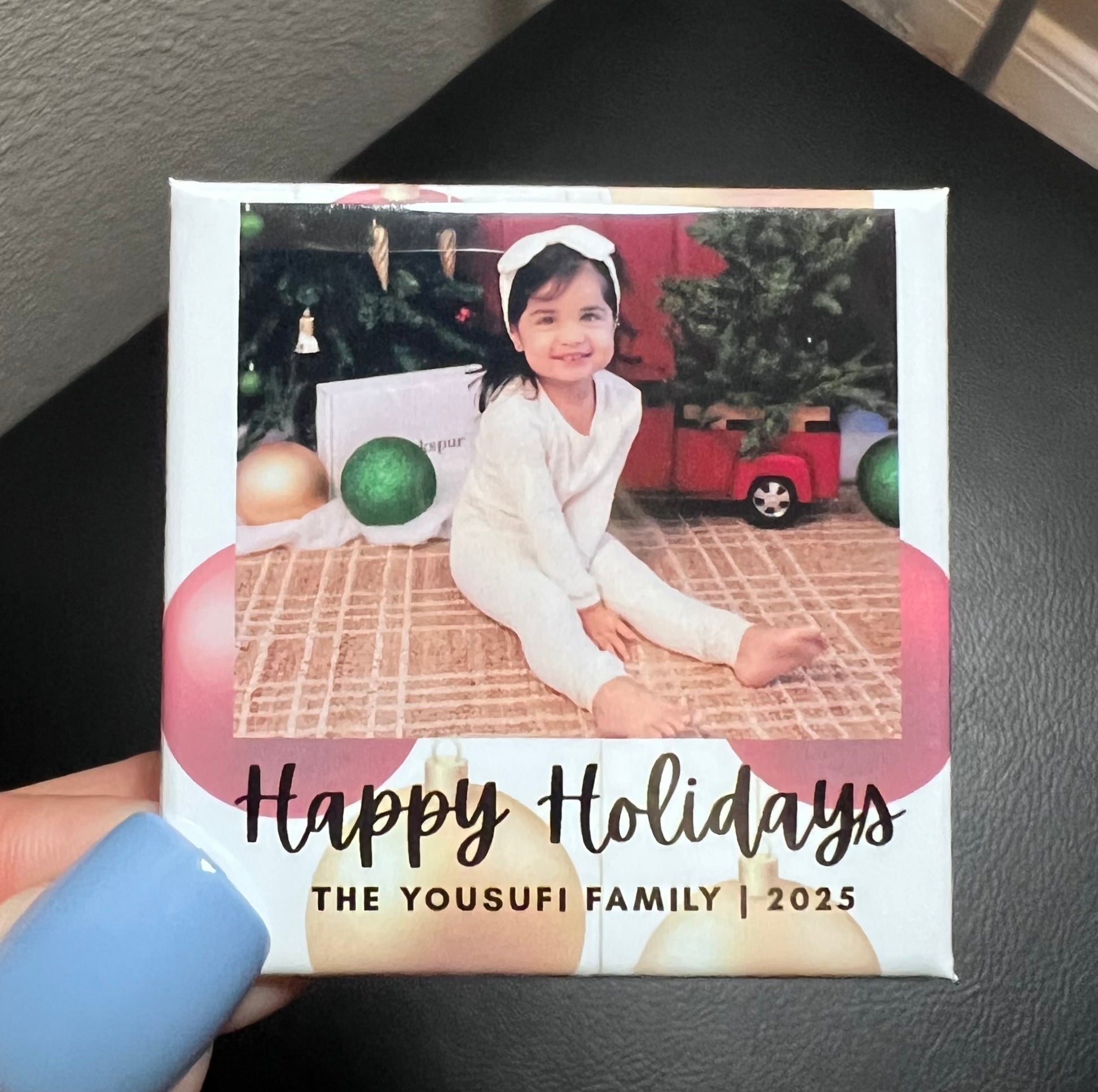 Holiday photo magnet with a child in front of a Christmas tree, held by a person's hand.
